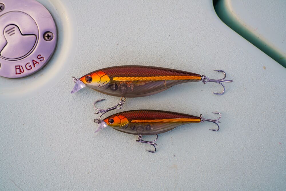 Two fishing crankbait lures with a metallic bronze-brown body, bright orange and yellow accents, and purple treble hooks, resting on a light-colored boat surface next to a gas cap.