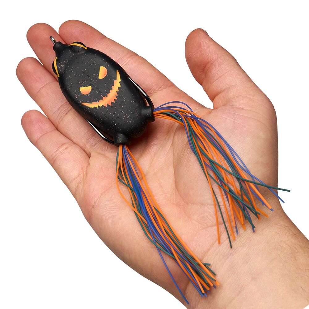 A hand holding a black pumpkin-shaped fishing lure with a glowing orange jack-o'-lantern face and multicolored dangling strands in orange, blue, and green, against a plain white background.