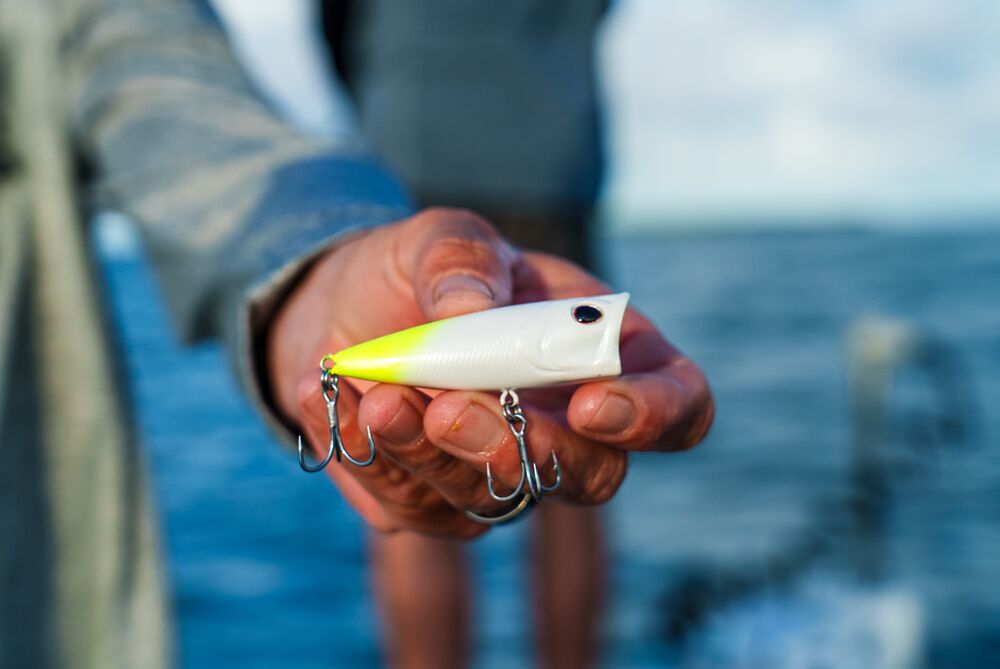 A close-up of a person's hand holding a white and neon yellow fishing lure with three silver treble hooks, set against a blurred background of blue water and sky.