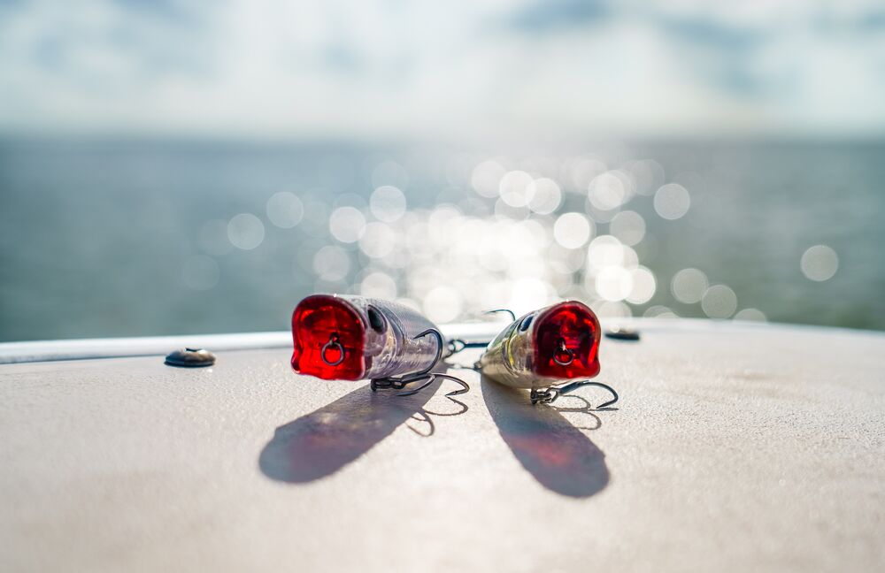 Two fishing lures with red reflective heads and silver bodies resting on a boat deck, with a blurred ocean and sky background.