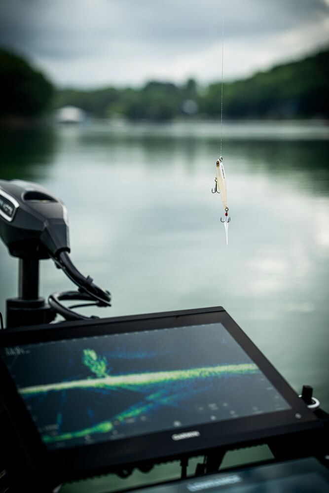 A close-up of a fishing lure on a line in the foreground, with a boat's display screen and motor mount visible, set against a blurred lake and tree shoreline.