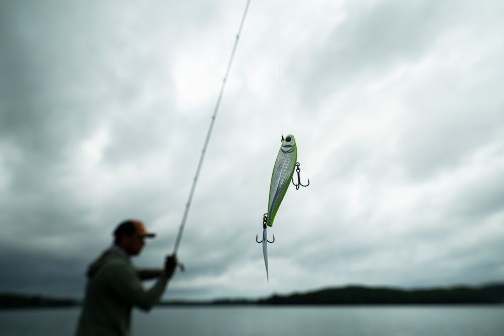 A fishing lure shaped like a small silver and green fish is suspended mid-air from a rod held by a blurred fisherman on a lake under a cloudy gray sky.