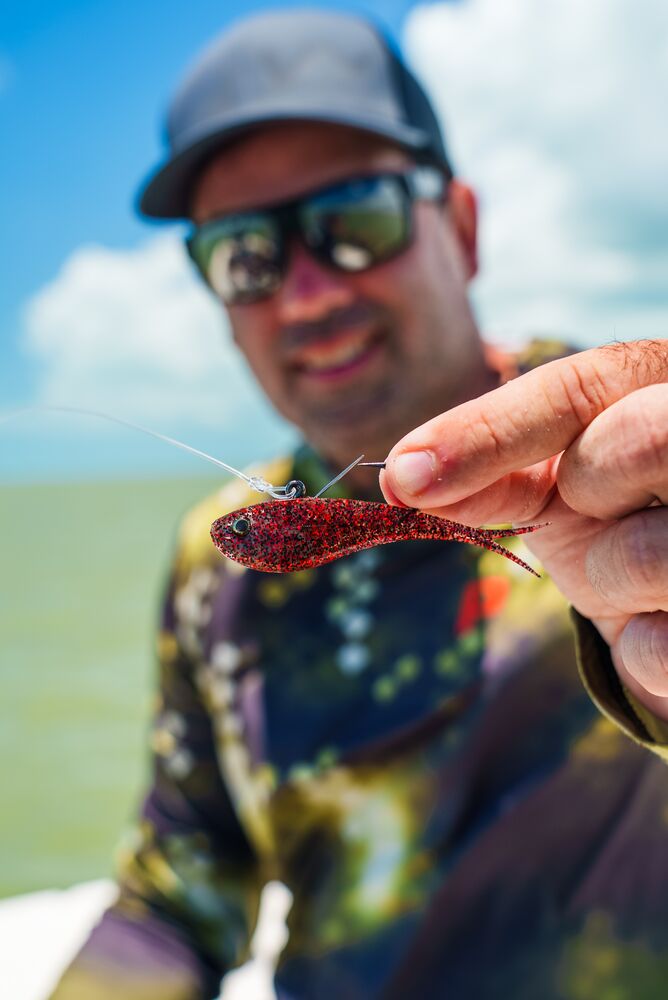 A close-up lifestyle shot of a fisherman holding a small red and black soft plastic fishing lure in front of his face. The fisherman is wearing a camouflage long-sleeve shirt, sunglasses, and a dark cap, with a blurred outdoor background of blue sky and water.