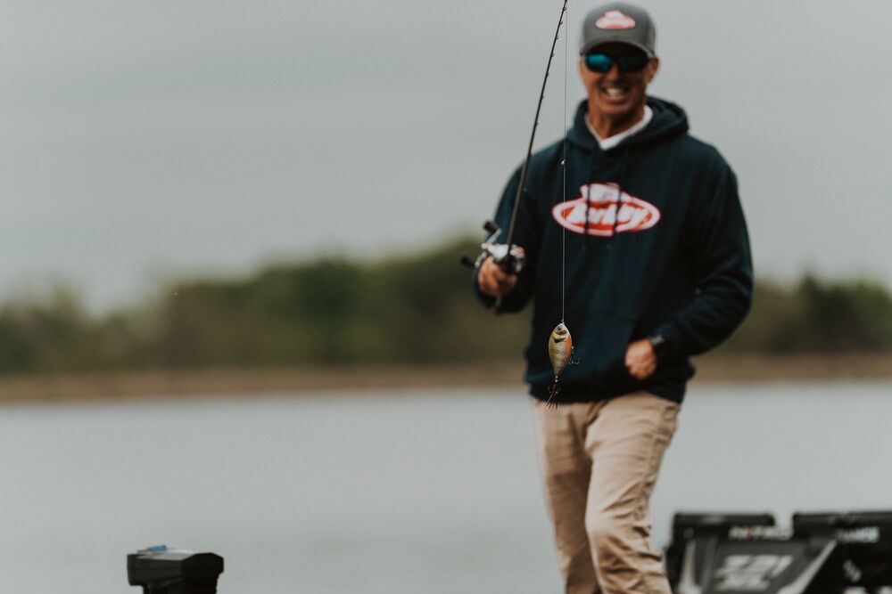A man wearing a dark navy hoodie with a red and white logo, khaki pants, and a cap stands on a boat holding a fishing rod and lure over calm water.