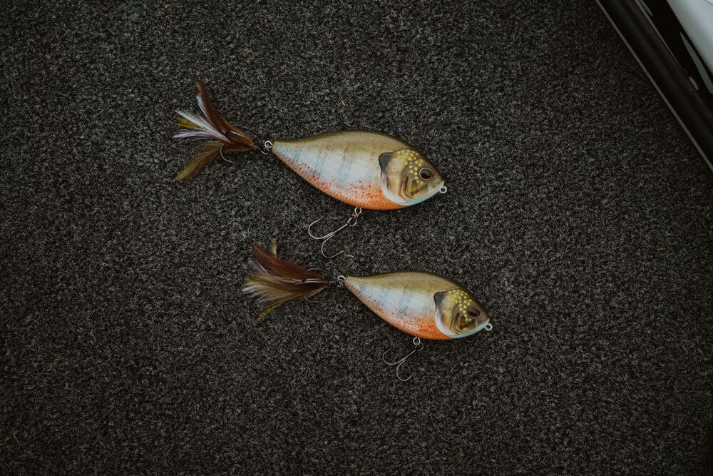 Two artificial fishing lures shaped like small fish, placed on a dark textured carpet. Each lure has a silver and orange body with a gold head and brown-and-white feather tail.