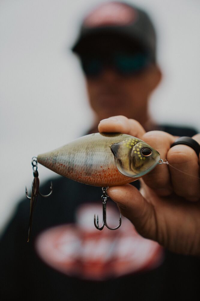 A close-up of a person holding a realistic fishing lure shaped like a small fish, with two treble hooks attached. The person is blurred in the background, wearing a dark cap and shirt.